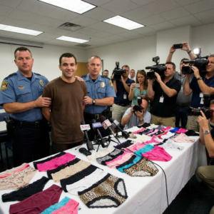 In a brightly lit indoor setting, likely a press conference room, two uniformed police officers flank a man in a plain brown t-shirt. The officers, wearing blue shirts with epaulets and badges, have a firm but neutral grip on the man's shoulders, directing him towards the cameras. The man [image uploaded] with short dark hair, is smiling subtly and maintaining eye contact with the lens.

Stretching across the foreground is a white-draped table, upon which a diverse collection of women's underwear is meticulously displayed. There are dozens of pairs in various colors, patterns, and styles, including solid hues of red, blue, pink, and black, as well as leopard print and other designs. Several microphones with news station logos are placed among the undergarments, pointing towards the man and officers.

Behind the table, a throng of journalists and photographers are actively documenting the event. Many are holding professional video cameras with large lenses and attached lights, while others are using DSLR cameras with flash attachments, and some are capturing footage with their smartphones. The room is illuminated by rectangular fluorescent light panels on the ceiling, casting an even glow. The overall atmosphere suggests a formal public presentation, possibly a police press briefing or an arrest announcement.