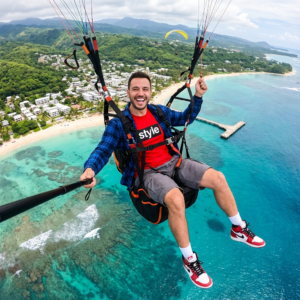 Ultra-realistic, vibrant aerial adventure photograph captures a joyful man (same face as reference image, 100% accuracy) paragliding high above a breathtaking tropical coastline. He is seated comfortably in his black and orange harness, legs dangling freely, facing the camera with a wide, excited smile, teeth showing, conveying pure exhilaration. He sports dark brown hair and a light beard,wearing a bright red t-shirt emblazoned with 'style' in bold white text within a blackCteate a Ultra-realistic, vibrant aerial adventure photograph in 9:16 ratio captures a joyful man (same face as reference image, 100% accuracy) paragliding high above a breathtaking tropical coastline. He is seated comfortably in his black and orange harness, legs dangling freely, facing the camera with a wide, excited smile, teeth showing, conveying pure exhilaration. He sports dark brown hair and a light beard,wearing a bright red t-shirt emblazoned with 'style' in bold white text within a blackrectangle, layered beneath an open blue and black plaid long-sleeved shirt. His attire includes grey cargo shorts, white socks, and distinctive red, white, and black high-top sneakers. In his left hand, he holds a black selfie stick, while his right grasps the paragliding control lines. The stunning backdrop features crystal-clear turquoise and deep blue ocean revealing intricate coral reefs and gentle breaking waves near a pristine white sandy beach. A charming coastal town with numerouswhite buildings is nestled against lush, emerald-green, tree-covered hills, which ascend to distant, hazy mountains under a softly clouded sky. Another yellow paraglider gracefully soars in the distance, and a long concrete pier extends into the tranquil water. The scene is illuminated by bright, diffused natural light, creating an exhilarating, free-spirited, and idyllic tropical mood, with a color palette dominated by vivid turquoises, deep blues, and verdant greens, beautifully contrastedby the crisp whites of the beach and town, and the bold reds of his clothing and footwear.