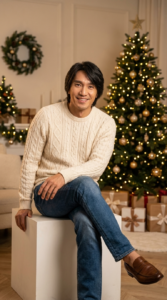 Create a hyper-realistic 8K close-up body portrait of a female model, using the uploaded photo as the exact facial reference. Maintain 100% accuracy of the facial features — do not alter or modify any aspect of the face. Render the skin texture, lighting, and overall composition with photo-realistic detail, ensuring lifelike color tones and natural depth of field. A man with his original hair is sitting casually on a white cube, smiling warmly at the camera. He is wearing a cream-colored cable-knit sweater, blue jeans, and brown loafers. His legs are crossed, with one hand resting on his knee.
The background reveals a cozy and festive living room. A large, beautifully decorated Christmas tree with numerous warm lights and gold ornaments stands prominently behind him. Several wrapped gift boxes are visible at the base of the tree. To his left, another smaller decorated Christmas tree and a wreath on the wall further enhance the holiday atmosphere. The lighting is soft and inviting, creating a warm and welcoming scene.