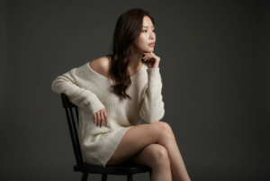 Generate an ultra-realistic, highly ultra-detailed, 8k resolution with 1080x1080 pixel portrait of me using the uploaded image for reference (preserved the likeness and the original face for reference) of a striking, high-resolution portrait of a young woman with long, flowing wavy brunette hair, seated gracefully on a simple black wooden chair in a dimly lit studio. She wears an oversized, off-white knitted sweater that drapes loosely over her frame, slipping off one shoulder to reveal smooth skin and add a subtle touch of sensuality. Her pose is elegant and contemplative: one arm rests gently on the chair’s backrest while her hand delicately supports her chin, and her gaze is directed thoughtfully toward the side, creating an intimate and introspective mood. The lighting is expertly crafted with a single soft, directional light source positioned to the side, casting gentle shadows that sculpt her form and highlight the texture of the knitwear and the natural contours of her legs and arms. This chiaroscuro effect enhances the depth and dimensionality of the image, emphasizing the softness of her skin and the intricate weave of the sweater. The background is a smooth, muted dark gray, providing a minimalist and distraction-free backdrop that contrasts beautifully with her light-colored apparel and warm skin tones. *** The composition is a full-body vertical frame that captures the model’s seated posture with perfect balance, positioning her slightly off-center to create visual interest and harmony. The overall color palette is subdued and warm, featuring neutral tones that evoke a sense of calm and understated elegance. The image is impeccably sharp, showcasing fine details such as the delicate waves of her hair, the subtle folds in the fabric, and the natural texture of her skin. The style is classic and timeless, blending modern minimalism with emotive portraiture to convey quiet strength, beauty, and introspection.