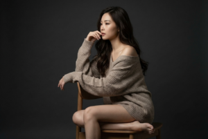 Generate an ultra-realistic, highly ultra-detailed, 8k resolution with 1080x1080 pixel portrait of me using the uploaded image for reference (preserved the likeness and the original face for reference) of a cinematic studio portrait of a woman seated on a simple wooden chair with a minimalist design, positioned slightly to the left of the frame. She is captured in a contemplative pose, with her body turned to the left, her left arm resting gracefully on the back of the chair, and her right hand gently touching her face near her lips, conveying a sense of introspection and elegance. Her long, wavy hair cascades naturally over her shoulders, framing her face and adding softness to the composition. She wears an oversized, textured knit sweater that slips off her shoulders, exposing her collarbones and upper chest, emphasizing a relaxed and intimate mood. Her legs are bare, with her right foot flat on the ground and her left knee slightly raised, creating a dynamic line that guides the viewer’s eye through the composition. *** The background is a seamless, deep charcoal or dark brown studio backdrop, providing a rich, neutral setting that enhances the dramatic lighting. The lighting setup features a single, soft yet directional light source positioned to the left of the subject, casting gentle, sculptural shadows that highlight the contours of her face, shoulders, and arms, while creating a subtle gradient across her form. The light accentuates the texture of her sweater and the natural shine of her hair, adding depth and dimension to the image. The color palette is monochromatic with warm, muted tones—shades of gray, brown, and beige—contributing to a timeless, artistic aesthetic. The image is shot with a professional full-frame camera using an 85mm or 50mm lens at a wide aperture (f/1.8 to f/2.😎 to achieve a shallow depth of field, ensuring the subject is in sharp focus while the background remains softly blurred. The resolution is ultra-high, capturing every detail from the fine texture of her sweater to the subtle expression of her pose. The overall style is elegant, contemplative, and refined, emphasizing mood and atmosphere over overt glamour. Post-processing is minimal, maintaining natural skin tones, enhancing contrast and clarity, and preserving the authenticity of the scene. This portrait embodies a delicate balance between simplicity and emotional depth, making it suitable for fine art, editorial, or fashion photography.