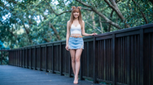 一張年輕東方女性站在戶外木棧道上的全身人像照片。
人物與穿著:
這位女性擁有修長的身材和淺棕色的長直髮，留著齊劉海。
她面帶微笑，表情甜美。頭上戴著一對動物耳朵的髮飾。
上半身穿著一件白色的短版 T 恤/緊身衣（Crop Top），領口較低。
下半身是一條淺藍色的牛仔短裙，裙襬帶有蕾絲邊緣。繫著一條淺棕色的皮帶
腳上穿著一雙露趾的裸色高跟涼鞋。
背景與環境:
她站在一條戶外的深色木質棧道上，左手輕輕扶著旁邊的深色木質欄杆。
背景呈現柔和的散景（景深很淺），可以看到綠色的植物和一些被處理成偏冷藍色調的樹枝或樹葉，營造出一種清新且夢幻的氛圍。
總體而言，這張照片風格清新、甜美，光線柔和，突出了人物的時尚穿搭和外貌。