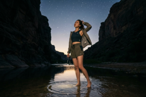 Base Setup
keep 100 percent facial information adherence of the attached image and turn her into a lone night hiker standing in the shallow river at the mouth of the canyon, captured as a live action photograph or movie still, not an illustration or CGI render, with an alluring, confident, quietly sexy mood.

Shot and Camera
Three quarter body shot from slightly low height on the riverbank, placing her on the left third while the water leads into the dark canyon and the star filled sky dominates the top of the frame, preserving the original wide vertical composition.

Identity and Pose
She has a slim athletic build, natural proportions, medium length loose hair, and lightly tanned skin, 8k Photorealistic and hyper realistic. She stands barefoot in the water with one leg forward, hips relaxed, wearing a fitted cropped technical top and separate high waisted trail shorts with an open lightweight jacket slipping off one shoulder, one hand loosely holding a headlamp at her side and the other brushing hair from her face as she looks up at the stars.

Lighting and Environment
Keep the soft starlight and faint glow from the distant horizon, giving a gentle rim on her shoulders and hair while a subtle fill from the reflected sky reveals her features. The canyon walls, river reflections, pebbled shore, and dense star field stay exactly like the reference, with small ripples around her feet grounding her in the water.

Masking and Constraints
Change only by adding the subject and her outfit, keep lighting, perspective, white balance, canyon geometry, and river reflections the same, with realistic scale, body proportions, and clear contact between feet and water surface. Change only the requested element and keep lighting, perspective, white balance, pose, face geometry, body proportions, and silhouette the same, and absolutely avoid added text, painterly or toon styling, CGI look, blown highlights, banding, plastic skin, overly tidy backgrounds, or floating feet while requiring consistent perspective, consistent white balance, subtle film like grain, and physically correct contact shadows and reflections.
