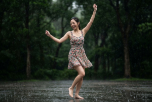 Edit the uploaded photo (face based on the reference photo). Ensure the face remains consistent with the person in the uploaded image, without changing facial structure, skin tone . Create a Create an 8K ultra-realistic image of a jpyful woman dancing and celebrating in the rain outdoors. She has a big smile on her face, eyes closed with happiness. She is wearing a floral sleeveless dress with a fitted waist and a short, flowy skirt. She has a simple gold necklace and her dark hair is tied back. Her arms are raised, one hand higher than the other, and she is barefoot, standing on wet ground with her toes touching the surface. The background is blurred with dark green trees, emphasizing her joyful expression and movement. The rain is falling steadily around her, creating a lively and vibrant atmosphere. Using a Canon EOS R camera with a 50mm f/1.8 lens, f/2.2 aperture, shutter speed 1/200s, ISO 100 and natural light, Full Body, Hyper Realistic Photography, Cinematic, Cinema, Hyper detail, Ultra hd, Color Correction, ultra hd, hdr , color grading, 8k.