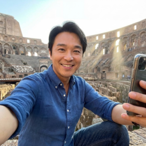 A selfie taken inside the Roman Colosseum. Insert {reference_image} as the face. He wears a button-down shirt and jeans, smiling as he holds the phone up. Sunbeams shine through the arches behind him. The face is sharp, centered, and well lit.