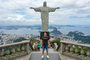 Drone photo near Christ the Redeemer in Rio. Include a close-up of the tourist using {reference_image} as the face, standing at the viewpoint. He wears a casual t-shirt, shorts, and sneakers. Drone hovers slightly in front and above him, capturing both his face clearly and the giant statue towering behind.