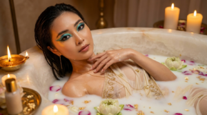 {
"intro": "Create an ultra realistic 8K UHD DSLR photo based on the attached image as a reference of facial features, maintaining 100% likeness.",

"subject": {
"identity": "A stylish beautiful woman portrayed as Cleopatra during her sacred milk bath beauty ritual, embodying divine femininity, sensual elegance, and timeless power.",
"angle": "Close-up beauty editorial captured at a refined 3/4 angle, focusing on her face, shoulders, and upper chest with extreme clarity and no blur.",
"pose": {
"body_position": "She is partially submerged in a luxurious milk bath, her shoulders and collarbones emerging gracefully from the surface.",
"hands": "One hand gently rests at the edge of the bath with milk droplets on her fingers, while the other lightly touches her neck adorned with subtle gold jewelry.",
"expression": "Soft yet commanding gaze directed toward the camera—calm, confident, intimate, and hypnotic."
}
},

"appearance": {
"outfit": "A barely-there, ritual-style ivory silk drape partially submerged in the milk bath, clinging softly to her skin. The fabric is delicate and translucent, edged with fine gold-thread embroidery for a sensual, sacred aesthetic.",
"accessories": "Minimal ritual jewelry—thin gold collar necklace resting above the milk surface, delicate arm cuff, subtle finger ring—kept refined to maintain focus on beauty and skin.",
"hair": "Her hair is sleek and ritual-polished, partially damp and slicked back at the crown, with soft wet strands framing her face naturally, reflecting candlelight.",
"makeup": "High-fashion Cleopatra beauty makeup with ceremonial artistry—perfectly sculpted brows, elongated black kohl eyeliner, vibrant eyeshadow layered in turquoise, teal, emerald, sapphire blue, and metallic gold. Beneath the eyes, subtle artistic ink-inspired accents echo ancient Egyptian symbolism. Her skin is luminous and dewy, cheeks softly flushed, and lips finished in a nude-rose satin sheen."
},

"ritual_props": {
"bath": "A carved alabaster bathtub filled with warm milk infused with honey, almond oil, and lotus essence.",
"floating_elements": [
"Fresh lotus flowers",
"Soft white rose petals",
"Delicate gold flakes shimmering on the milk surface"
],
"beauty_products": [
"Small golden perfume vial with lotus and myrrh oil",
"Alabaster bowl of honey and milk mixture",
"Clay jar of mineral kohl and pigment powders"
]
},

"background": {
"macro_environment": "An intimate royal bathing chamber within Cleopatra’s palace, designed as a sacred beauty sanctuary.",
"midground_details": "Soft linen curtains, glowing oil lamps, tall candles, and subtle steam rising from the bath, creating a warm and sensual atmosphere.",
"micro_elements": "Milk ripples around her skin, floating petals touching her shoulders, tiny droplets of moisture on her collarbones, reflections of candle flames on gold jewelry, visible stone texture on the tub, and delicate fabric translucency—every detail sharply rendered with zero blur."
},

"lighting": {
"type": "Warm cinematic candlelight combined with soft ambient palace glow.",
"effect": "Golden highlights sculpt her face and skin while gentle shadows enhance depth, intimacy, and divine beauty."
},

"camera": {
"camera_type": "DSLR",
"resolution": "8K UHD",
"lens": "85mm prime lens for beauty editorial compression",
"aperture": "f/8 for maximum facial and detail sharpness",
"iso": 100,
"shutter_speed": "1/160s",
"focus": "Extreme sharp focus on facial features, skin texture, makeup details, and milk surface, no bokeh, no blur"
},

"style": "Luxury beauty editorial, sacred milk bath ritual, ancient Egyptian goddess realism, intimate yet powerful, ultra-detailed, luminous skin focus, cinematic elegance"
}