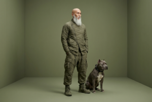 A full-body studio portrait of an elderly man with a bald head and a long, thick white beard, standing confidently beside a muscular grey pit bull dog. The man wears layered monochromatic olive-green tactical fashion: a quilted asymmetric jacket with mixed textures, loose padded cargo pants, and rugged military-style sneakers. His hands rest casually in his pockets as he gazes thoughtfully to the side, calm and powerful. The dog sits alert at his side, wearing a black studded collar, matching the strong, loyal aesthetic. Minimalist studio background in soft matte olive green, seamless floor and wall. Soft cinematic lighting with subtle shadows, high fashion editorial style, ultra-detailed fabric textures, muted earthy color palette, sharp focus, realistic skin tones, dramatic yet elegant mood, 8K resolution, professional fashion photography, Vogue editorial, modern urban warrior aesthetic.