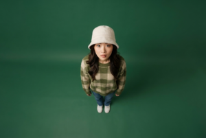 A cinematic top-down portrait of a young woman standing on a solid deep green floor, captured from an extreme overhead angle. She looks up directly at the camera with wide, expressive eyes, creating an intimate and slightly surreal mood. She wears a soft white bucket hat, a cozy oversized green-and-beige checkered sweater, blue jeans, and white sneakers. Minimalist composition with vast negative space surrounding her, emphasizing isolation and calm. Soft diffused studio lighting, natural skin tones, subtle shadows, clean color grading with earthy greens, editorial fashion photography style, ultra-sharp focus, high resolution, modern aesthetic, cinematic framing, shot on a professional DSLR, shallow depth of field, Instagram poster vibe